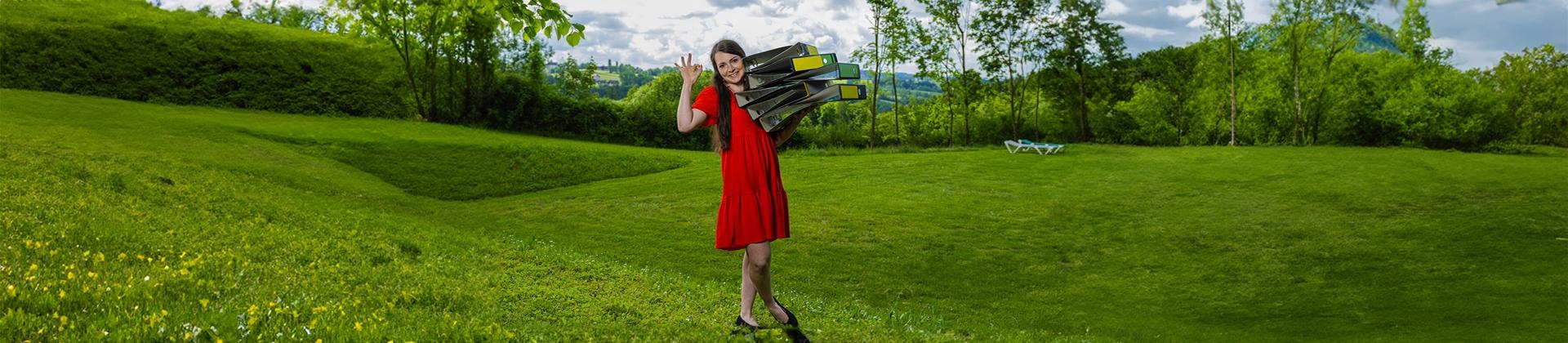 Frau mit Ordnern auf dem Arm steht mitten in einer grünen Hügellandschaft.